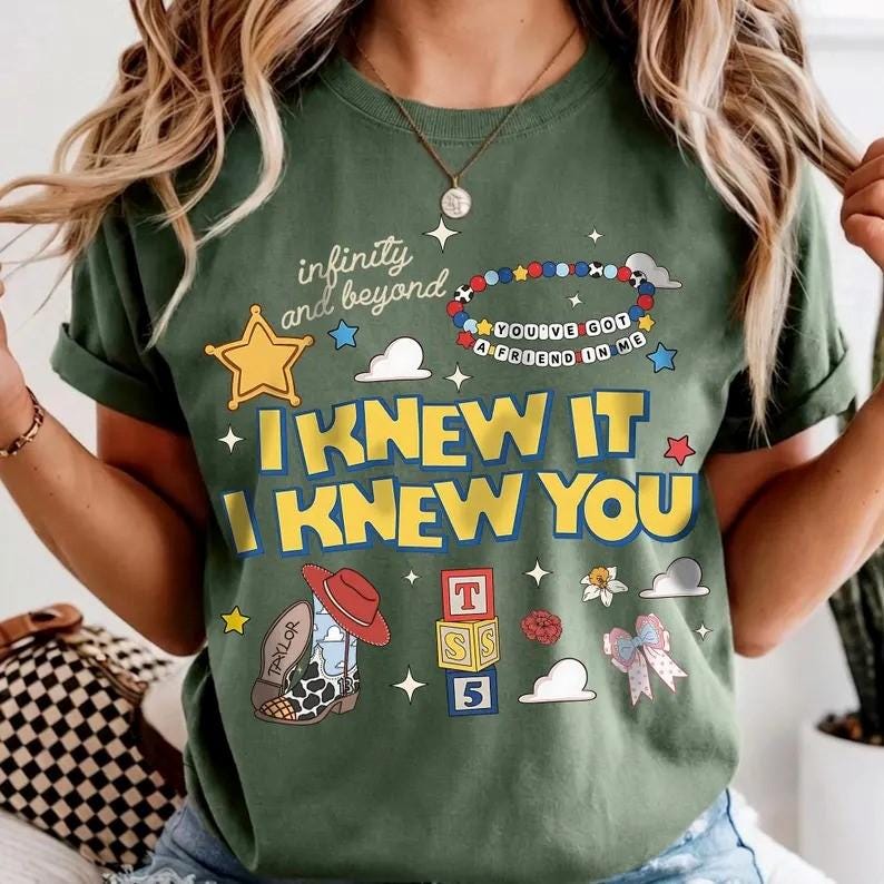 May include: An olive green t-shirt featuring the text "infinity and beyond", "I KNEW IT I KNEW YOU", and "YOU'VE GOT A FRIEND IN ME" on a beaded bracelet graphic. Additional illustrations include a yellow star, a cowboy boot, alphabet blocks, and a bow.