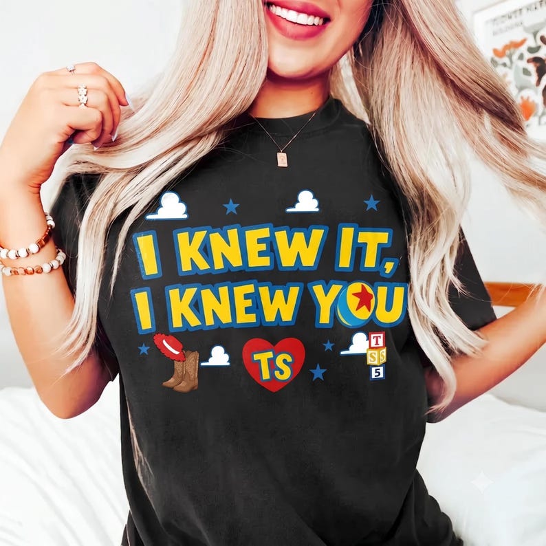 Op de afbeelding: Een zwart t-shirt met korte mouwen en de geel-blauwe tekst I KNEW IT, I KNEW YOU. Het ontwerp bevat een rood hart met de letters TS, een paar bruine cowboylaarzen met een rode hoed, een kleurrijke bal en gestapelde letterblokken.