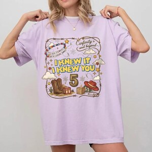May include: A light purple t-shirt featuring a graphic print with the text "Cinfinity and beyond" and "I KNEW IT I KNEW YOU". The design includes a large number 5, cowboy boots, a cowboy hat, alphabet blocks, stars, clouds, and a lasso border.