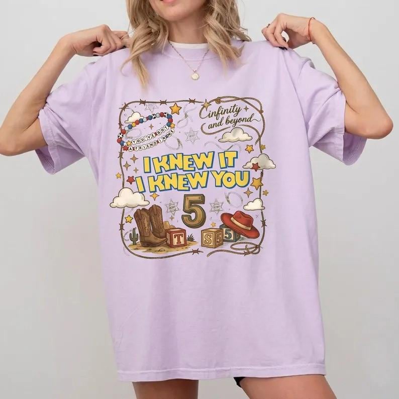 May include: A light purple t-shirt featuring a graphic print with the text "Cinfinity and beyond" and "I KNEW IT I KNEW YOU". The design includes a large number 5, cowboy boots, a cowboy hat, alphabet blocks, stars, clouds, and a lasso border.