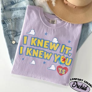 May include: A light purple t-shirt featuring the yellow and blue text I KNEW IT, I KNEW YOU, accompanied by white cloud graphics, blue stars, a colorful ball with a red star, and a red heart containing the yellow letters TS on the front.