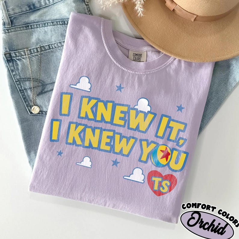 May include: A light purple t-shirt featuring the yellow and blue text I KNEW IT, I KNEW YOU, accompanied by white cloud graphics, blue stars, a colorful ball with a red star, and a red heart containing the yellow letters TS on the front.