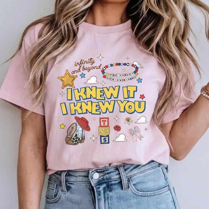 May include: A light pink t-shirt featuring a graphic print with the text "infinity and beyond," "I KNEW IT," "I KNEW YOU," and "YOU'VE GOT A FRIEND IN ME." The design includes a yellow star, a beaded bracelet, a cowboy boot, blocks, and a bow.