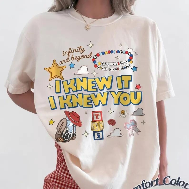 May include: A cream-colored short-sleeve t-shirt featuring a graphic print with the text "infinity and beyond," "YOU'VE GOT A FRIEND IN ME," and "I KNEW IT I KNEW YOU." The design includes a yellow star, a beaded bracelet, a cowboy boot, and blocks.