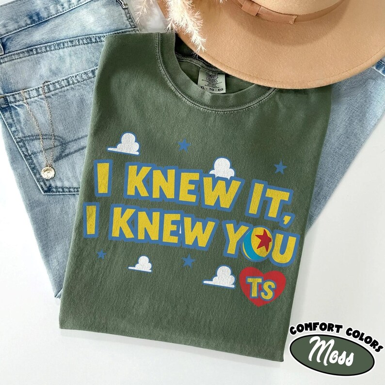 May include: An olive green t-shirt featuring the yellow text I KNEW IT, I KNEW YOU, a red heart with the yellow letters TS, a colorful ball graphic, and white cloud shapes. The shirt is displayed alongside light blue denim jeans and a tan hat.