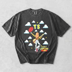 May include: A charcoal gray t-shirt featuring a graphic of a person with long hair wearing a yellow plaid shirt and blue jeans, holding a guitar. The design includes white clouds, yellow stars, a red hat, the text TS, and I KNEW IT I KNEW YOU.