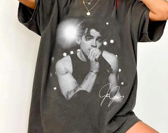 Op de afbeelding: Een antracietgrijs T-shirt met korte mouwen en een zwart-witte grafische print van een persoon die in een microfoon zingt. Het ontwerp bevat een helder lichteffect achter de figuur en een witte handgeschreven handtekening rechtsonder.