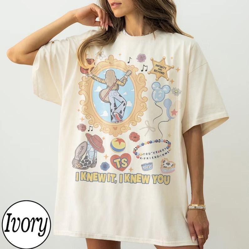 May include: An ivory short-sleeve t-shirt featuring a graphic print with a central ornate gold frame containing a figure with a guitar, surrounded by icons including a star with text Infinity and Beyond, balloons, a cowboy boot, and the text I KNEW IT, I KNEW YOU.