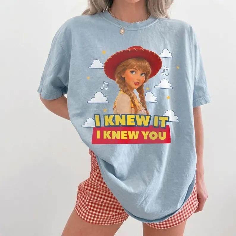 May include: A light blue t-shirt featuring a graphic of a character with braided hair wearing a red cowboy hat. The design includes white clouds, yellow stars, musical notes, and the text I KNEW IT I KNEW YOU in yellow and red block letters.