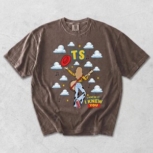 May include: A brown short-sleeve t-shirt featuring a graphic of a person with long hair wearing a yellow plaid shirt and cow-print pants, holding a guitar. The design includes white clouds, a red hat, yellow stars, and the text TS I KNEW IT I KNEW YOU.