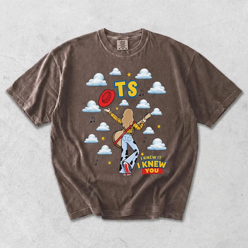 May include: A brown short-sleeve t-shirt featuring a graphic of a person with long hair wearing a yellow plaid shirt and cow-print pants, holding a guitar. The design includes white clouds, a red hat, yellow stars, and the text TS I KNEW IT I KNEW YOU.