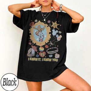 May include: A black t-shirt featuring a graphic print with a central ornate gold frame containing a figure with a guitar, surrounded by icons including a sheriff star, balloons, a cowboy boot, a beaded bracelet, and the text I KNEW IT, I KNEW YOU.