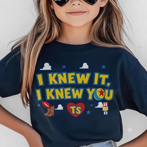 Op de afbeelding: Een donkerblauw t-shirt met de gele tekst "I KNEW IT, I KNEW YOU" en een rood hart met de letters "TS". Het ontwerp bevat een rode cowboyhoed, bruine laarzen, een kleurrijke bal, letterblokken en kleine blauwe sterren.