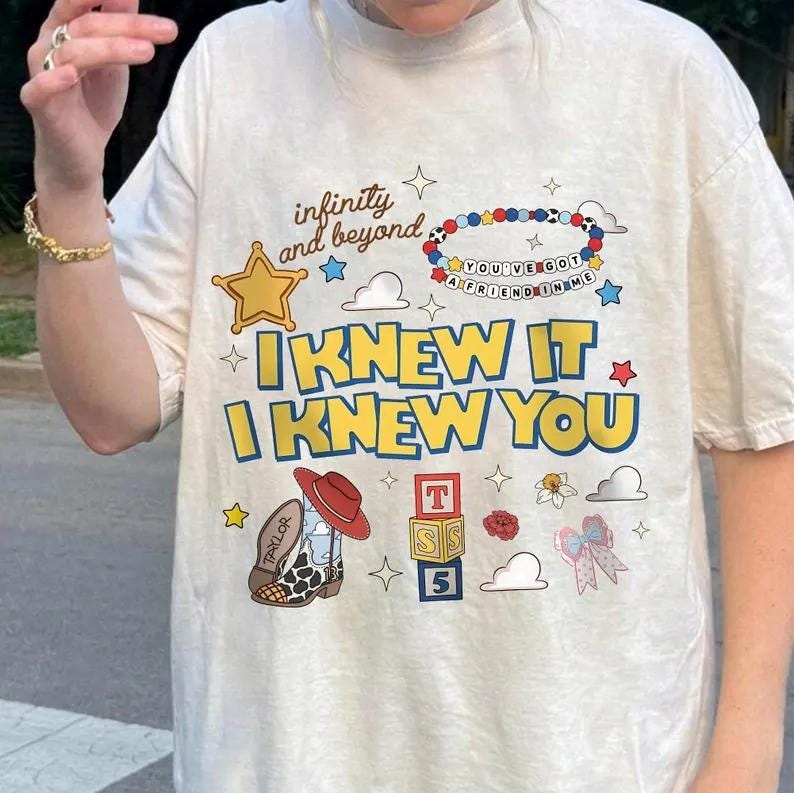 May include: An off-white short-sleeve t-shirt featuring a graphic print with the text "infinity and beyond," "YOU'VE GOT A FRIEND IN ME," and "I KNEW IT I KNEW YOU." The design includes a beaded bracelet, a cowboy boot, alphabet blocks, and stars.