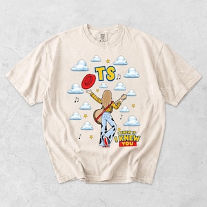 May include: A cream-colored t-shirt featuring a graphic of a person with long hair wearing a yellow plaid shirt and cow-print bell-bottom pants, holding a guitar and a red hat. The design includes blue clouds, yellow stars, musical notes, and the text TS and I KNEW IT I KNEW YOU.