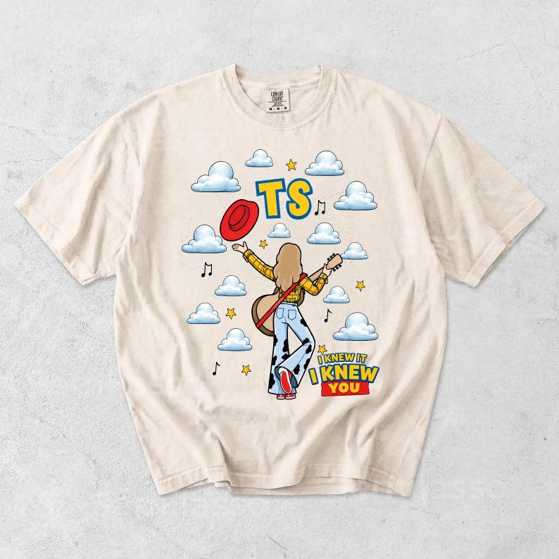 May include: A cream-colored t-shirt featuring a graphic of a person with long hair wearing a yellow plaid shirt and cow-print bell-bottom pants, holding a guitar and a red hat. The design includes blue clouds, yellow stars, musical notes, and the text TS and I KNEW IT I KNEW YOU.