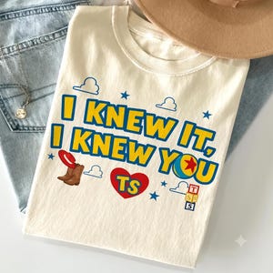 Op de afbeelding: Een cr&egrave;mekleurig t-shirt met de blauwe en gele tekst I KNEW IT, I KNEW YOU. Het ontwerp bevat een rode cowboyhoed, een paar bruine laarzen, een rood hart met de letters TS, een kleurrijke bal en een stapel van drie blokken met letters.