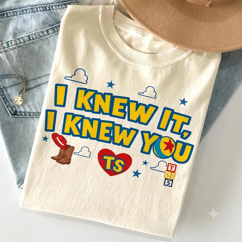 Op de afbeelding: Een cr&egrave;mekleurig t-shirt met de blauwe en gele tekst I KNEW IT, I KNEW YOU. Het ontwerp bevat een rode cowboyhoed, een paar bruine laarzen, een rood hart met de letters TS, een kleurrijke bal en een stapel van drie blokken met letters.