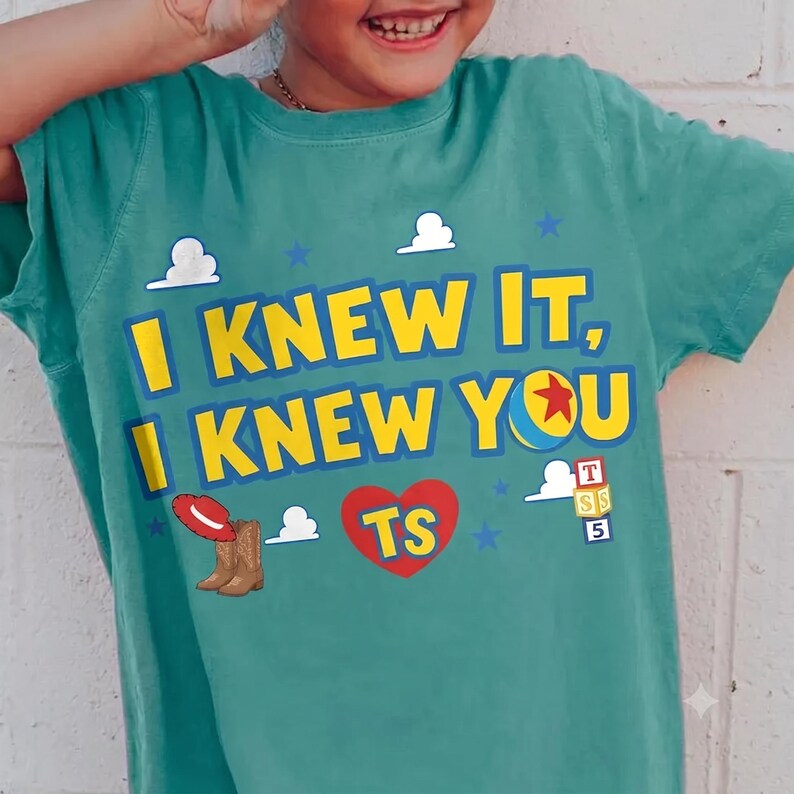 Op de afbeelding: Een blauwgroen t-shirt met korte mouwen en de gele tekst I KNEW IT, I KNEW YOU. Het ontwerp bevat een rode cowboyhoed, bruine laarzen, een rood hart met de letters TS, een kleurrijke bal, kleine witte wolken, blauwe sterren en blokken.