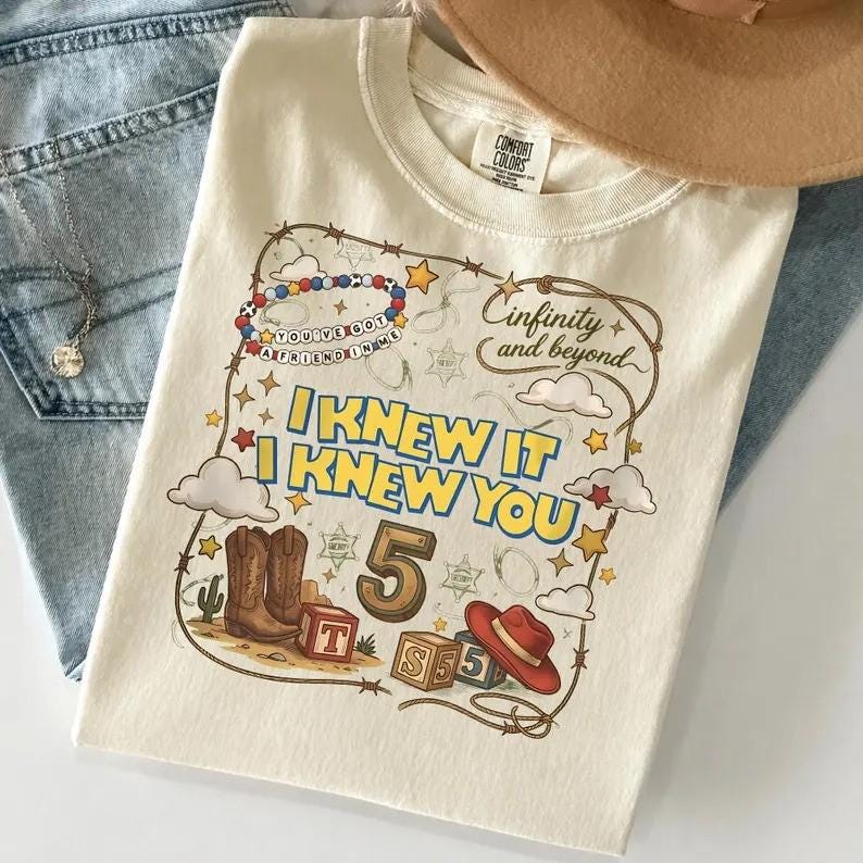 May include: A cream-colored t-shirt featuring a graphic print with the text "I KNEW IT I KNEW YOU", "infinity and beyond", and "YOU'VE GOT A FRIEND IN ME". The design includes cowboy boots, a red hat, alphabet blocks, stars, and clouds.