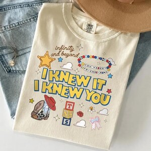 May include: A cream-colored t-shirt featuring a graphic print with the text "infinity and beyond," "YOU'VE GOT A FRIEND IN ME," and "I KNEW IT I KNEW YOU." The design includes a yellow star, a beaded bracelet, a cowboy boot, alphabet blocks, and clouds.