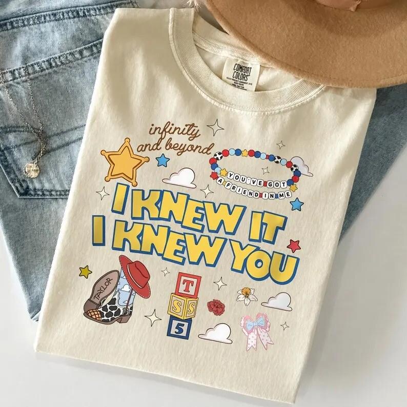 May include: A cream-colored t-shirt featuring a graphic print with the text "infinity and beyond," "YOU'VE GOT A FRIEND IN ME," and "I KNEW IT I KNEW YOU." The design includes a yellow star, a beaded bracelet, a cowboy boot, alphabet blocks, and clouds.