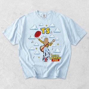 May include: A light blue t-shirt featuring a graphic of a person with long hair wearing a yellow plaid shirt and cow-print pants, holding a guitar and a red hat. The design includes clouds, stars, musical notes, and the text TS I KNEW IT I KNEW YOU.