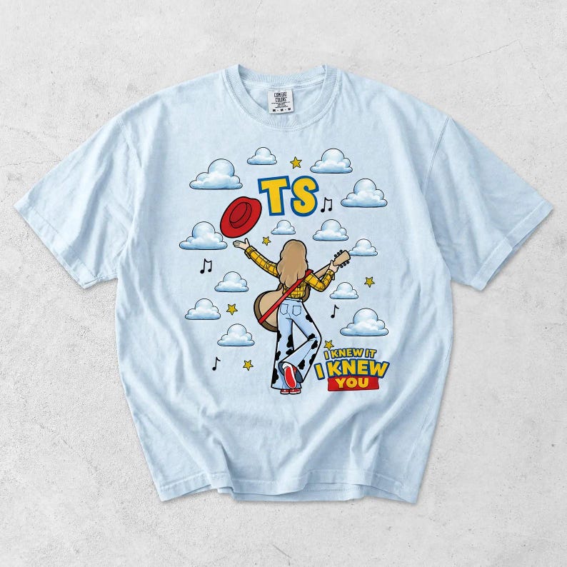 May include: A light blue t-shirt featuring a graphic of a person with long hair wearing a yellow plaid shirt and cow-print pants, holding a guitar and a red hat. The design includes clouds, stars, musical notes, and the text TS I KNEW IT I KNEW YOU.