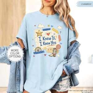 Puede incluir: Una camiseta de manga corta azul claro con un estampado gr&aacute;fico de una p&aacute;gina de bloc de notas rayada con el texto I Knew It, I Knew You, una estrella de sheriff, una bota vaquera, una rueda de carreta, un coraz&oacute;n y estrellas y nubes.