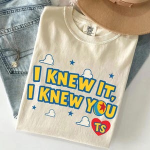 May include: A cream-coloured t-shirt featuring the blue and yellow text I KNEW IT, I KNEW YOU, with a red heart containing the letters TS, a colourful ball graphic, and small blue cloud and star icons printed on the front of the garment.
