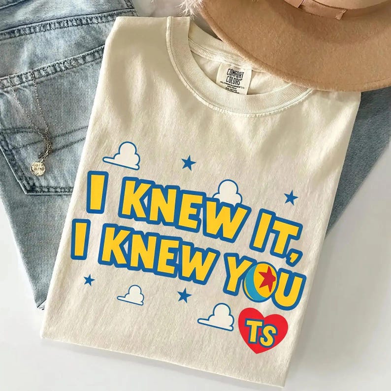 May include: A cream-coloured t-shirt featuring the blue and yellow text I KNEW IT, I KNEW YOU, with a red heart containing the letters TS, a colourful ball graphic, and small blue cloud and star icons printed on the front of the garment.