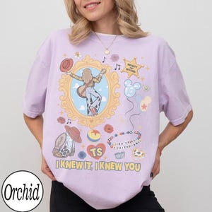 May include: A light purple t-shirt featuring a graphic print of a person with a guitar inside a gold frame, a cowboy boot, a ball, balloons, and the text "INFINITY AND BEYOND", "TS", "YOU'VE GOT A FRIEND IN ME", and "I KNEW IT, I KNEW YOU".