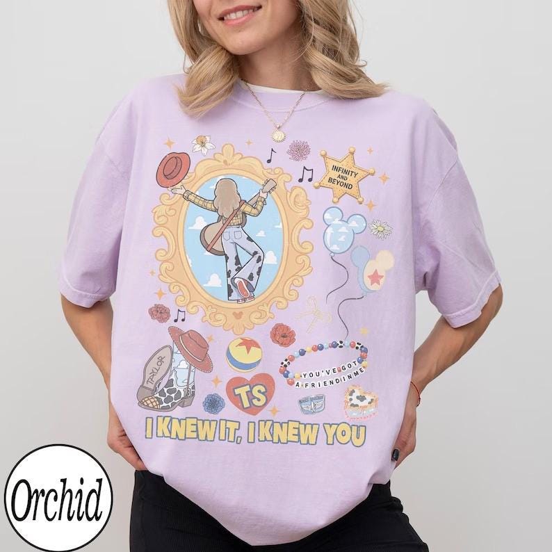 May include: A light purple t-shirt featuring a graphic print of a person with a guitar inside a gold frame, a cowboy boot, a ball, balloons, and the text "INFINITY AND BEYOND", "TS", "YOU'VE GOT A FRIEND IN ME", and "I KNEW IT, I KNEW YOU".