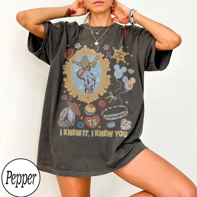 May include: A charcoal gray short-sleeve t-shirt featuring a graphic print with a central ornate gold frame containing a figure with a guitar. Surrounding elements include a star, balloons, a cowboy boot, and text reading I KNEW IT, I KNEW YOU.