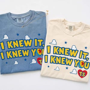 May include: Two short-sleeve crew neck t-shirts, one blue and one cream, each featuring the yellow text I KNEW IT, I KNEW YOU with a colourful ball graphic replacing the letter O, small white clouds, blue stars, and a red heart with the letters TS.