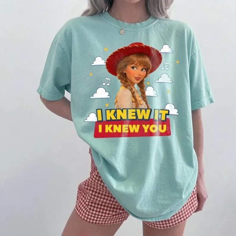 May include: A light mint green short-sleeve t-shirt featuring a graphic of a cartoon cowgirl with red hat and braids. The design includes white clouds, yellow stars, musical notes, and the text I KNEW IT I KNEW YOU in yellow and white letters.