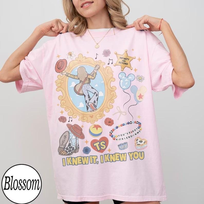 May include: A light pink t-shirt featuring a graphic print of a person with a guitar inside an ornate gold frame, surrounded by illustrations of a cowboy hat, boots, a ball, balloons, and text that reads "INFINITY AND BEYOND", "TS", and "I KNEW IT, I KNEW YOU".