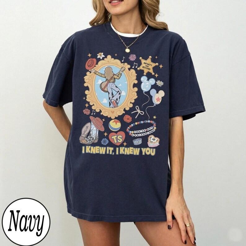 May include: A navy blue short-sleeve t-shirt featuring a graphic print of a gold ornate frame containing a person with a guitar, surrounded by icons including a star, balloons, a cowboy boot, and a ball. The text reads "INFINITY AND BEYOND" and "I KNEW IT, I KNEW YOU".