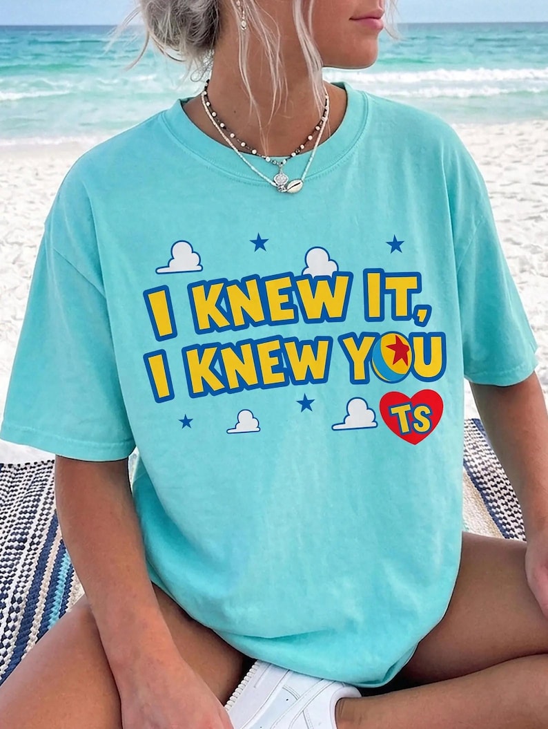May include: A light blue t-shirt featuring the yellow text I KNEW IT, I KNEW YOU with a red heart containing the letters TS. The design includes small white clouds and dark blue stars, with the letter O replaced by a yellow, red, and blue ball.