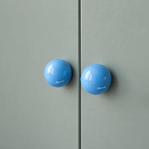 May include: Two spherical, glossy light blue cabinet knobs mounted on a sage green surface. Each knob features the white script text Ryland printed on the lower portion of its smooth, rounded surface, providing a clear contrast.