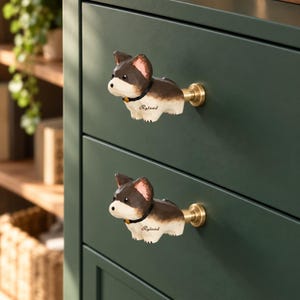 May include: Two drawer knobs shaped like a brown and white dog with a black collar. Each knob features the name Ryland printed in black script on the side and is attached to the dark green drawer front with a circular brass-toned metal base.