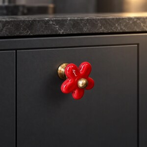 May include: A bright red, flower-shaped cabinet knob with a glossy finish and a gold-toned metal center. The word Ryland is printed in white script on one of the petals. The knob is mounted on a circular gold-toned metal base.