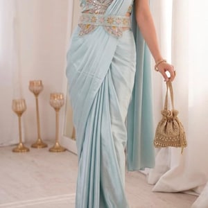 Puede incluir: Un sari azul claro con un top y cinturón a juego, con intrincados bordados y adornos. El conjunto incluye un pequeño bolso con cordón dorado. El sari está elegantemente drapeado, creando pliegues suaves y una silueta fluida.