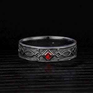 May include: A dark silver-toned metal ring featuring a textured band with an intricate, repeating geometric pattern. A single, small, square-cut red gemstone is set centrally within the band, serving as a focal point against the dark metal finish.
