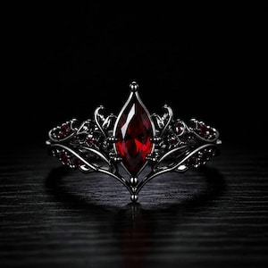 Può includere: Un anello in metallo nero con una gemma rossa centrale dal taglio marquise. La fascia è caratterizzata da un intricato lavoro di filigrana metallica a forma di viticcio che si estende verso l'esterno, con piccole pietre rosse incastonate.