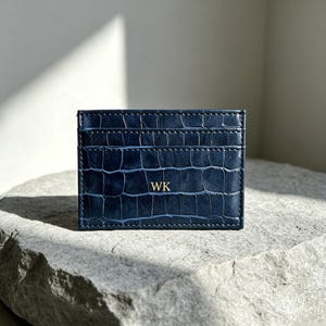 May include: A rectangular navy blue card holder featuring a textured crocodile skin pattern. The front surface displays the gold embossed initials WK centred in the middle, with visible horizontal stitching along the top and middle sections.