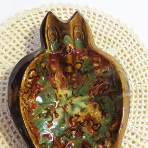 May include: A ceramic owl-shaped dish with a glossy finish. The dish features a brown exterior and a colorful interior with green, red, and yellow hues. The owl has a detailed design and sits on a cream-colored, crocheted doily.