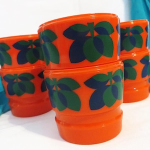 May include: A stack of orange plastic cups with a repeating pattern of green and blue leaf-like shapes. The cups are cylindrical with a slightly flared base and rim. The background is white with a hint of blue fabric.