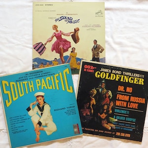May include: Three vintage vinyl record covers. The top cover is for "The Sound of Music" by Rodgers and Hammerstein. The middle cover is for "South Pacific" by Rodgers and Hammerstein. The bottom cover is for "Goldfinger" a James Bond thriller.