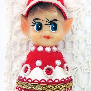 May include: A vintage Christmas ornament shaped like an elf. The elf has a red hat with a small green bell, blue eyes, and a red outfit decorated with pearls and red floral accents. The ornament is on a white lace background.